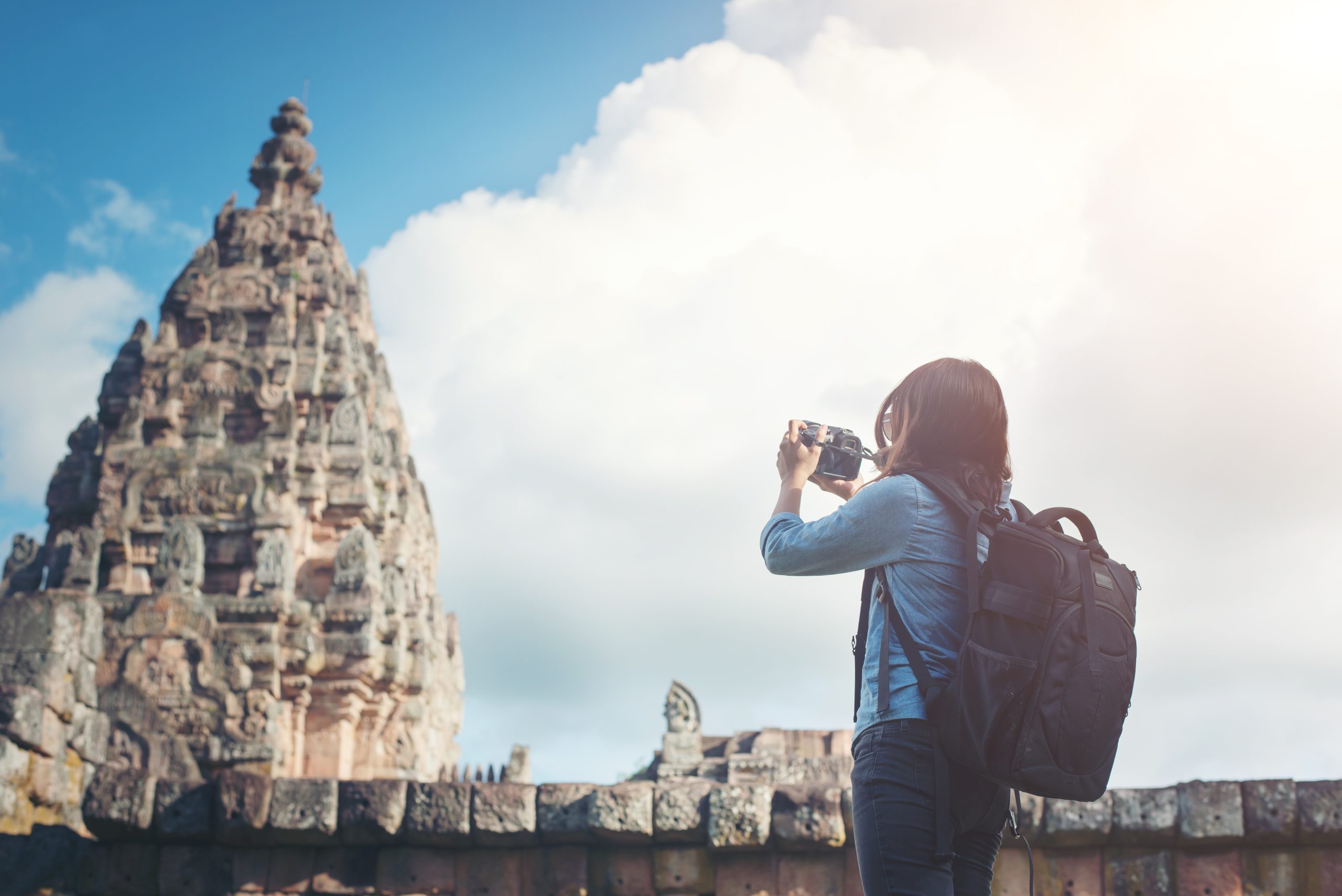 Female traveler with a backpack taking a photo of an ancient temple monument under a bright sky