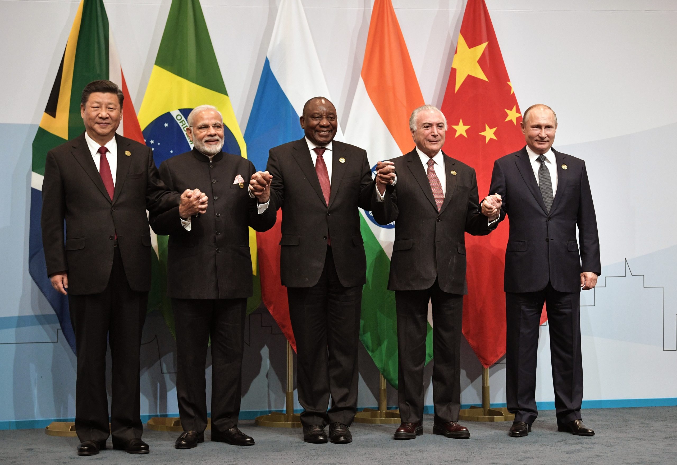 Leaders of BRICS nations standing together, holding hands in solidarity, with national flags of South Africa, Brazil, Russia, India, and China in the background.